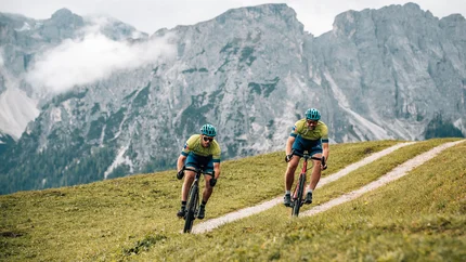 Herbst auf Schotter im Excelsior Dolomites Life Resort****s © Jens Scheibe Zwei Radfahrer fahren auf einem hügeligen Weg mit Bergen im Hintergrund