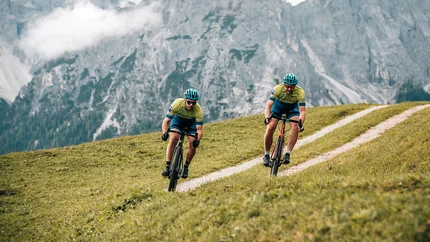 Two cyclists riding on a hilly path with mountains in the background