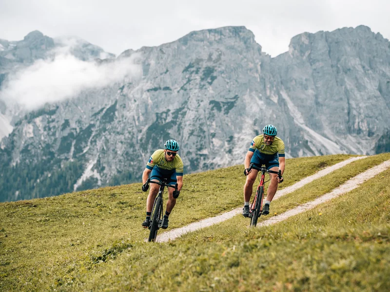 Gravelbike Holidays © Jens Scheibe Zwei Radfahrer fahren auf einem hügeligen Weg mit Bergen im Hintergrund