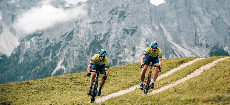 Zwei Radfahrer fahren auf einem hügeligen Weg mit Bergen im Hintergrund