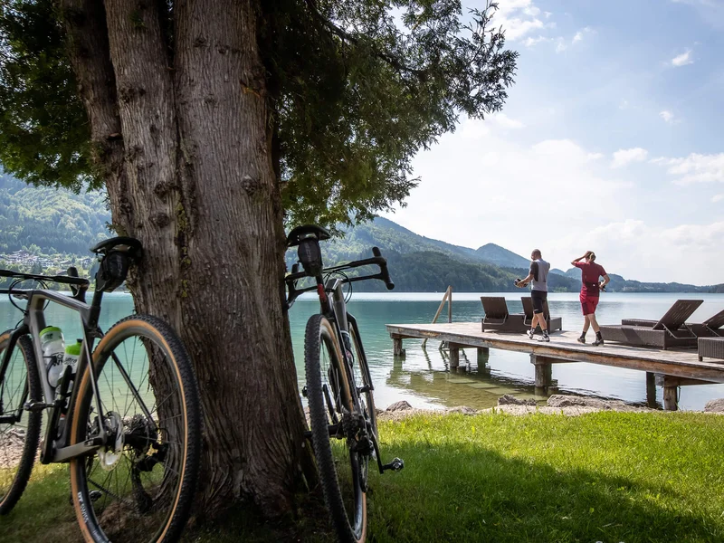 Gravelbike Holidays © Erwin Haiden Zwei Fahrräder lehnen an einem Baum am See mit Personen auf Steg