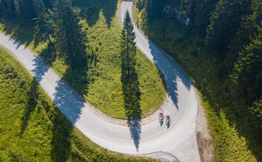 Gravelbike Urlaub in der Fuschlseeregion © Christina Neubauer Zwei Radfahrer auf einer kurvigen Bergstraße mit grünen Wiesen und Bäumen