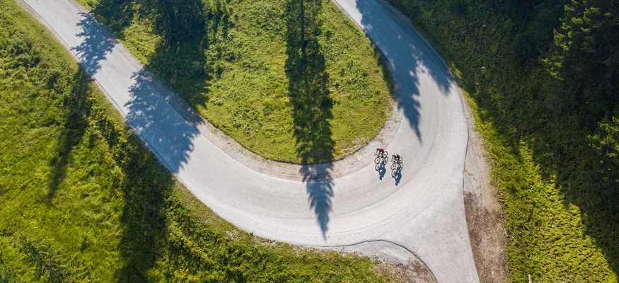 Two cyclists on a winding mountain road with green grass and trees