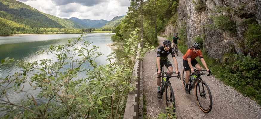 Drei Radfahrer fahren auf einem Pfad entlang eines Sees in bergiger Landschaft
