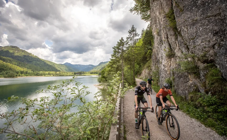 Three cyclists riding on a path beside a lake in mountainous scenery