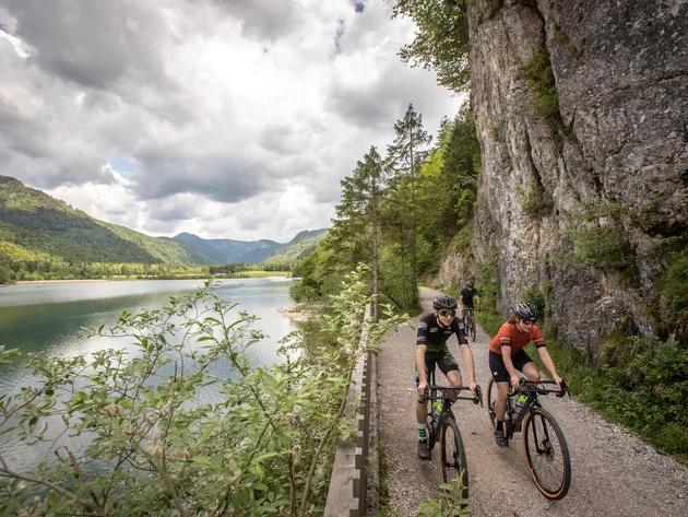 Drei Seen, eine Gravelrunde: Die 3-Seen-Tour ab Fuschl © Erwin Haiden Drei Radfahrer fahren auf einem Pfad entlang eines Sees in bergiger Landschaft