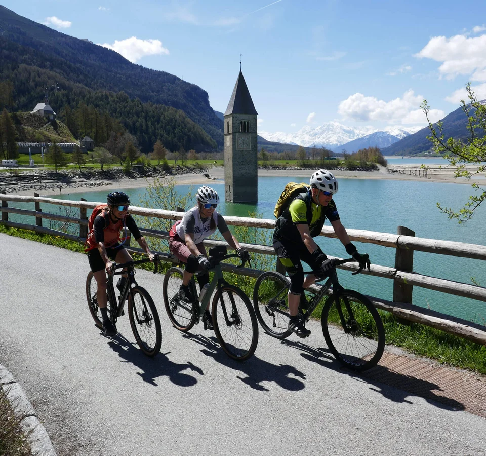 Drei Radfahrer fahren am Reschensee mit versunkenem Kirchturm im Hintergrund
