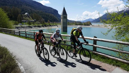 Drei Radfahrer fahren am Reschensee mit versunkenem Kirchturm im Hintergrund
