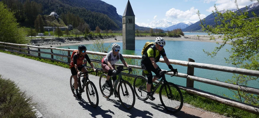 Drei Radfahrer fahren am Reschensee mit versunkenem Kirchturm im Hintergrund