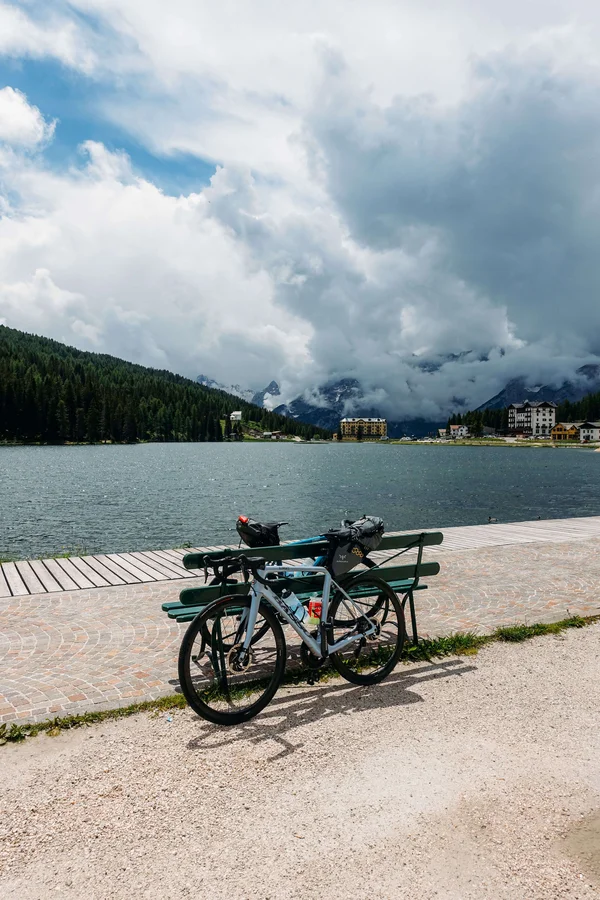 5 Gravelbike-Touren in Alta Badia, die du gefahren haben solltest © Hotel Melodia del Bosco Fahrrad an Bank am See mit bewölkten Bergen im Hintergrund