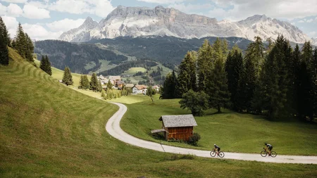 Two cyclists on a path in a green valley with mountains in the background