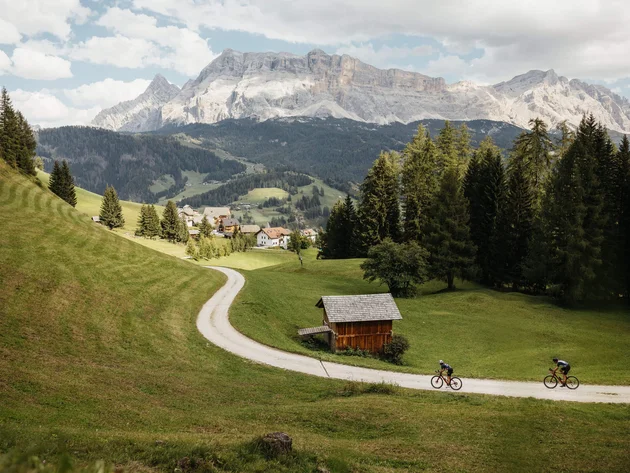 Two cyclists on a path in a green valley with mountains in the background