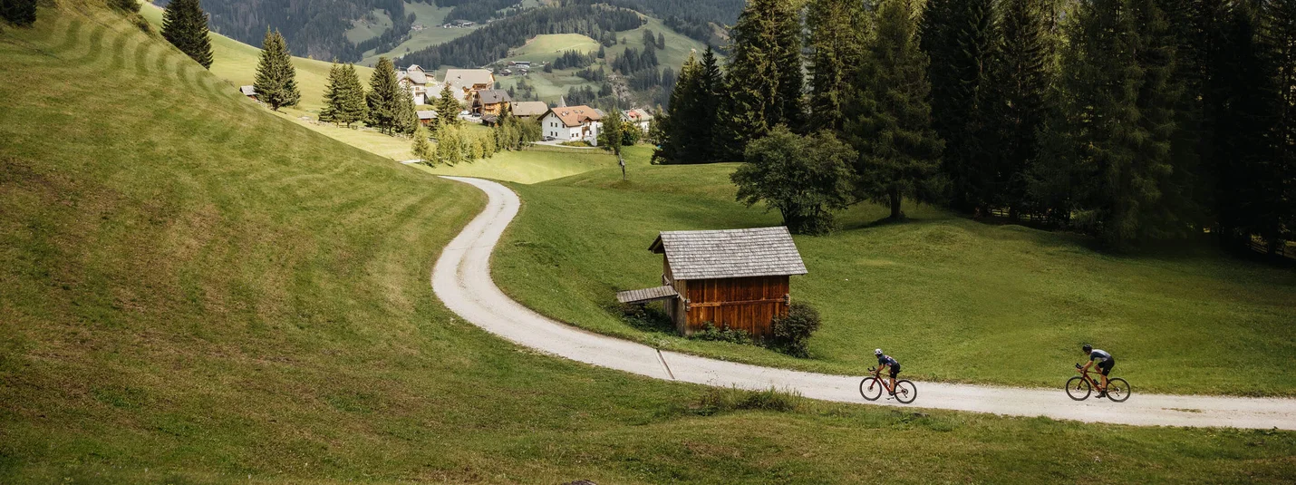 Gravel bike holiday in Alta Badia © Alex Moling Two cyclists on a path in a green valley with mountains in the background