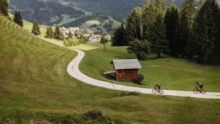 Two cyclists on a path in a green valley with mountains in the background