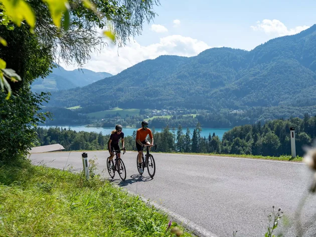 Drei Seen, eine Gravelrunde: Die 3-Seen-Tour ab Fuschl Zwei Radfahrer fahren auf einer Straße mit Berg- und Seeblick bei Sonnenschein