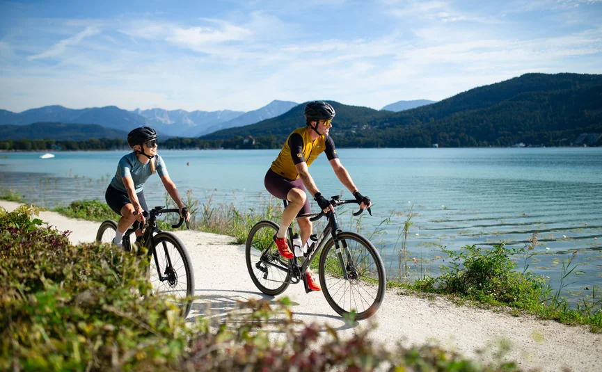 Two cyclists riding on a path beside a lake with mountains in the background