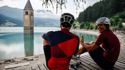 Zwei Radfahrer am See neben einem versunkenen Kirchturm sitzen und Eis essen
