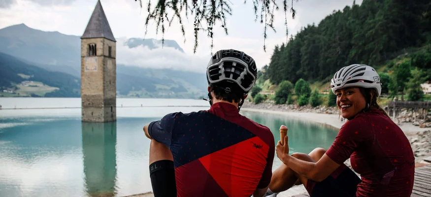 Zwei Radfahrer am See neben einem versunkenen Kirchturm sitzen und Eis essen