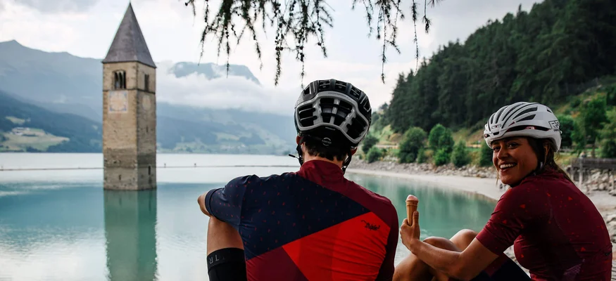 Zwei Radfahrer am See neben einem versunkenen Kirchturm sitzen und Eis essen