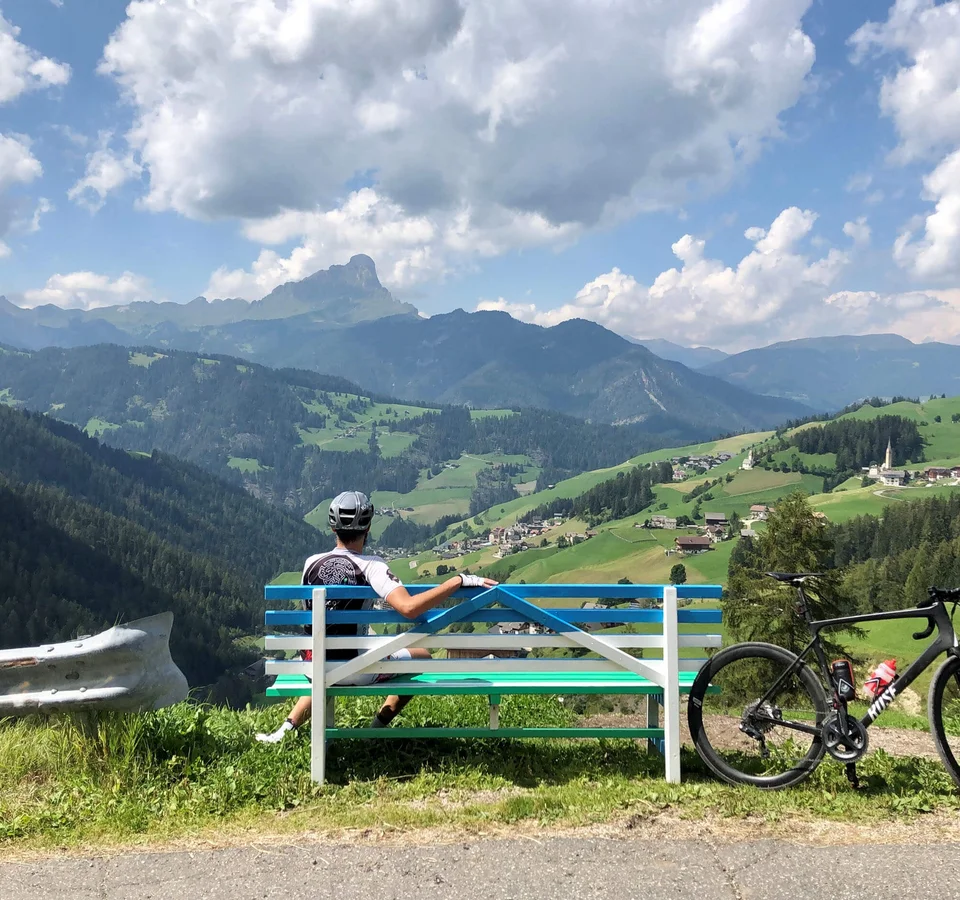 Gravel & Genuss am Kronplatz © Hotel Ostaria Posta Radfahrer auf Bank blickt auf grüne Berge und Dorf im Tal