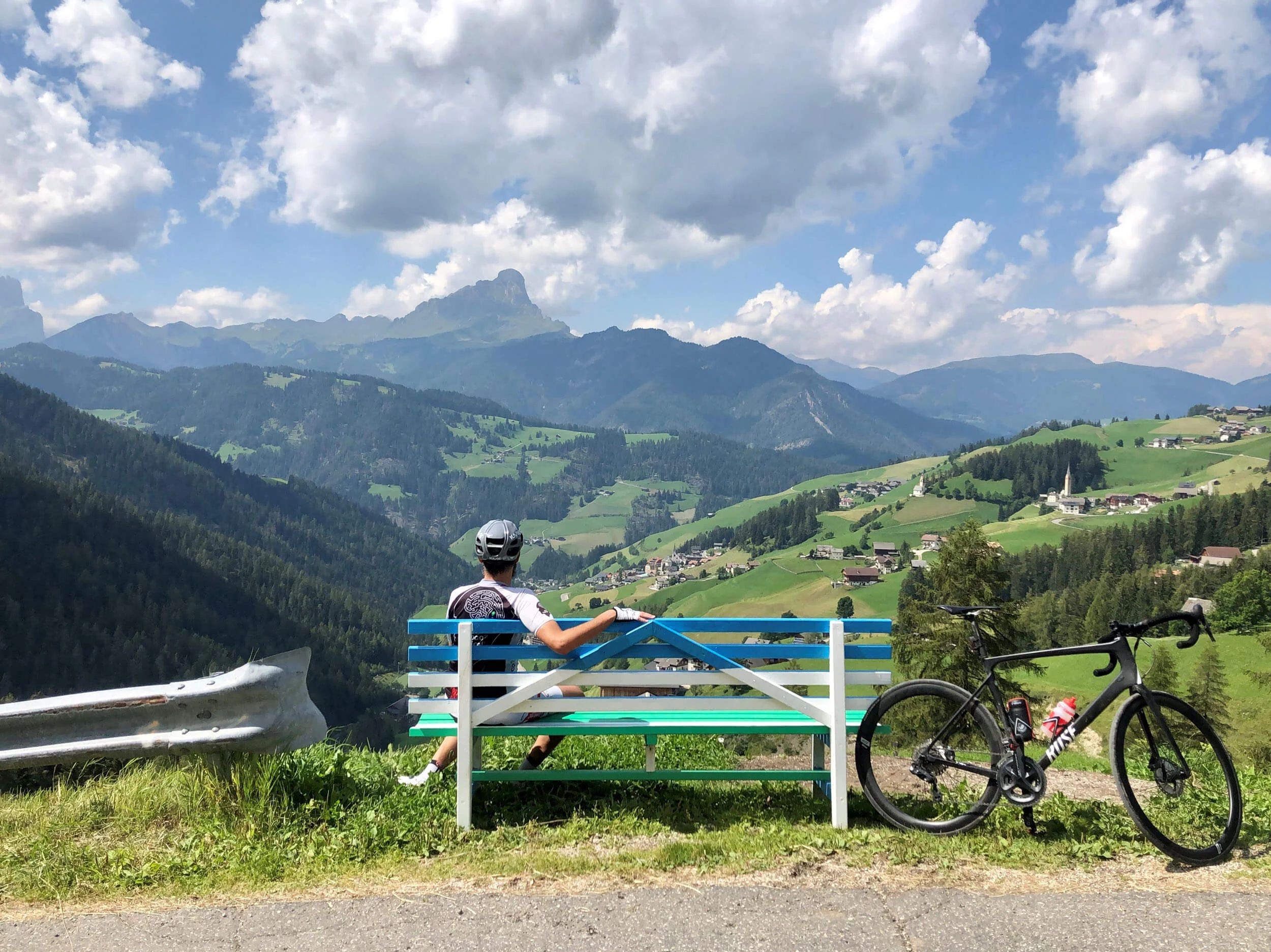 Epic Route Dolomites – 3 Zinnen – Gardasee © Hotel Ostaria Posta Radfahrer auf Bank blickt auf grüne Berge und Dorf im Tal