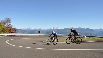 Zwei Radfahrer biegen auf kurviger Bergstraße mit Alpen im Hintergrund ab