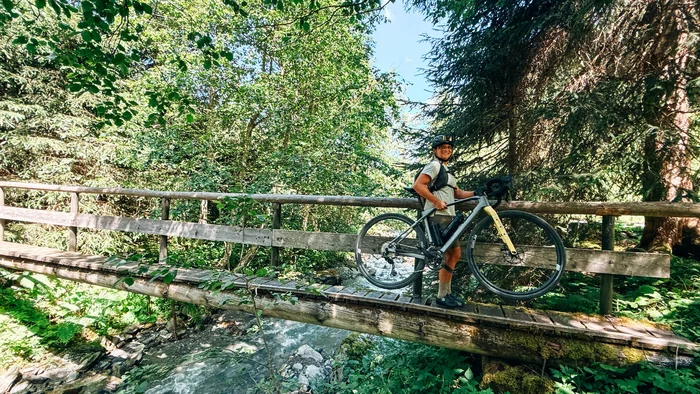 Graveln in den Kitzbüheler Alpen © Martina Wunderle / Flo Distl Person trägt Fahrrad über hölzerne Brücke in grünem Wald
