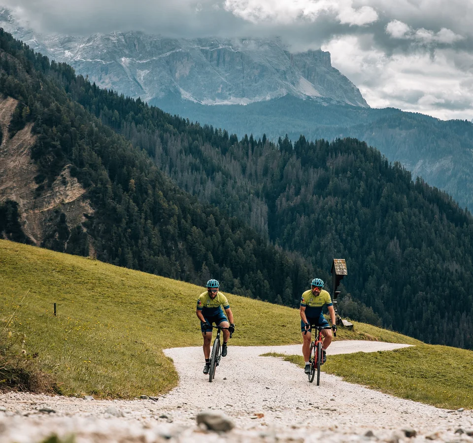 Zwei Radfahrer auf einem Bergweg vor bewaldeten Hügeln und bewölktem Himmel
