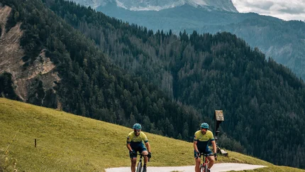 Zwei Radfahrer auf einem Bergweg vor bewaldeten Hügeln und bewölktem Himmel