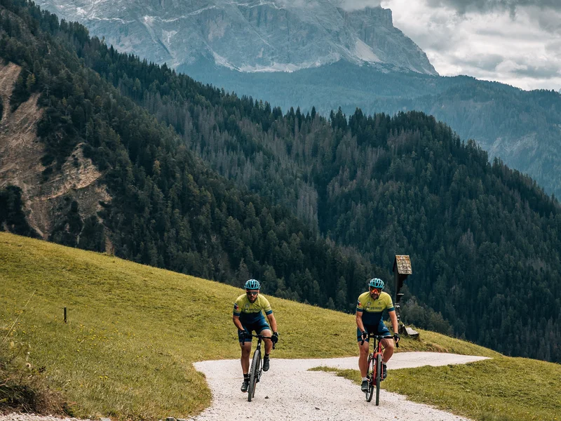 Zwei Radfahrer auf einem Bergweg vor bewaldeten Hügeln und bewölktem Himmel