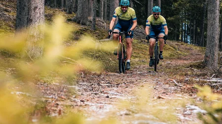 Herbst auf Schotter im Excelsior Dolomites Life Resort****s © Jens Scheibe Zwei Radfahrer fahren auf einem Waldweg zwischen Bäumen