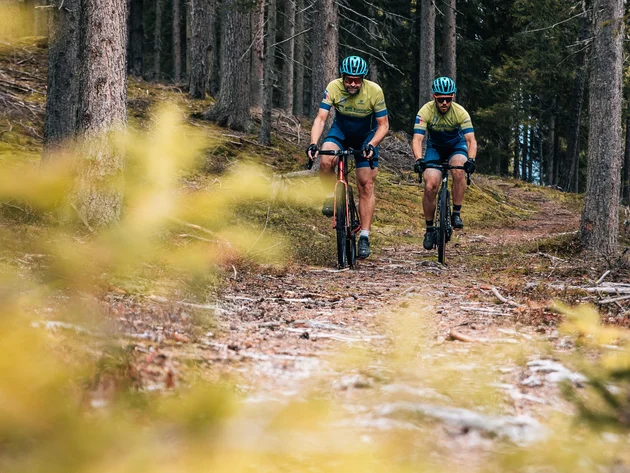 Herbst auf Schotter im Excelsior Dolomites Life Resort****s © Jens Scheibe Zwei Radfahrer fahren auf einem Waldweg zwischen Bäumen