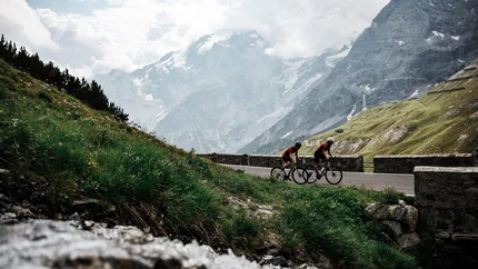 Two cyclists riding on mountain road with snow-covered peaks behind