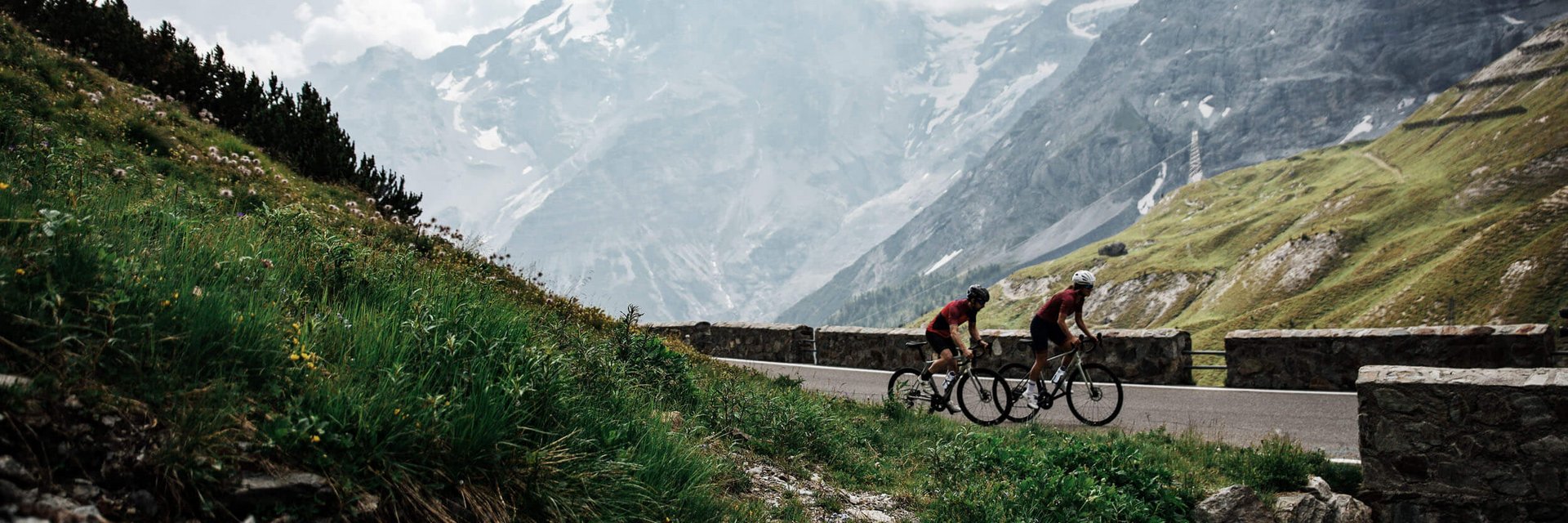 Two cyclists riding on mountain road with snow-covered peaks behind