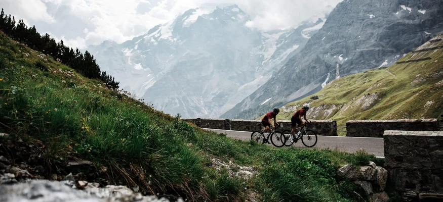 Zwei Radfahrer fahren auf Bergstraße mit schneebedeckten Bergen im Hintergrund