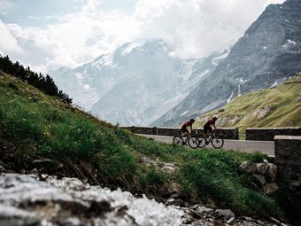 Zwei Radfahrer fahren auf Bergstraße mit schneebedeckten Bergen im Hintergrund