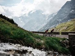 Two cyclists riding on mountain road with snow-covered peaks behind