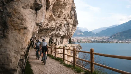 Radfahrer auf einem Pfad entlang einer Felswand mit Seeblick