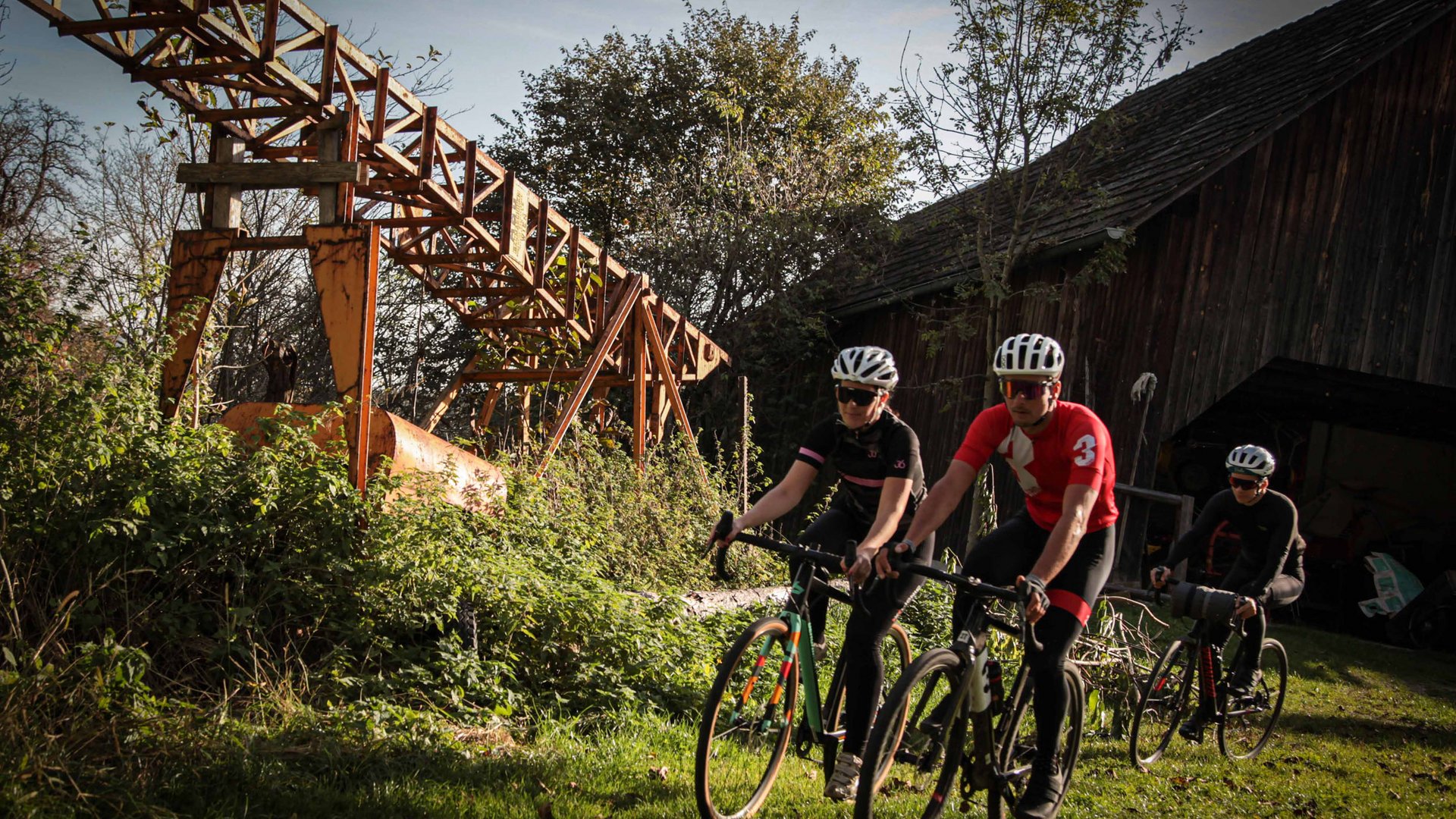 Drei Radfahrer fahren auf einem grasbewachsenen Weg an einer alten Industrieanlage vorbei