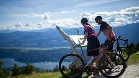Zwei Radfahrer lesen eine Tafel mit Bergblick und See im Hintergrund