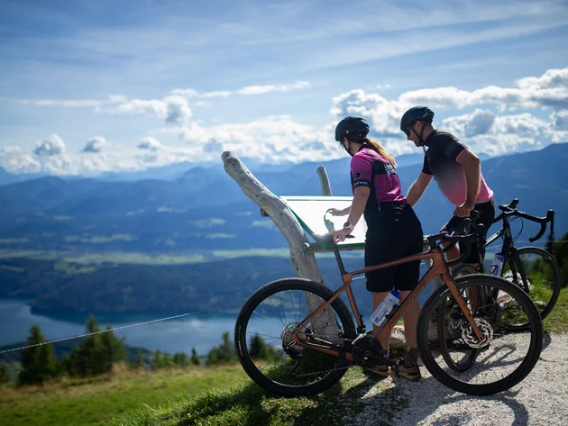 Zwei Radfahrer lesen eine Tafel mit Bergblick und See im Hintergrund