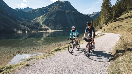 Zwei Radfahrer fahren auf einem Weg am See mit Bergen im Hintergrund