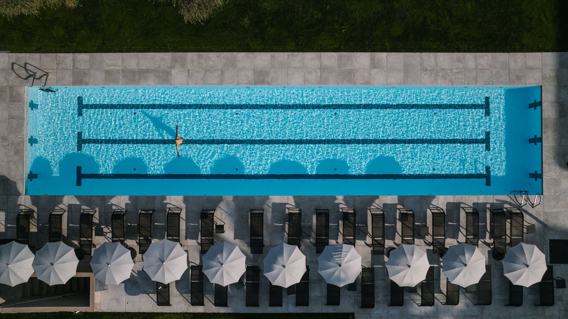Luftaufnahme eines Schwimmbads mit einer Person im Wasser und Liegestühlen mit Sonnenschirmen