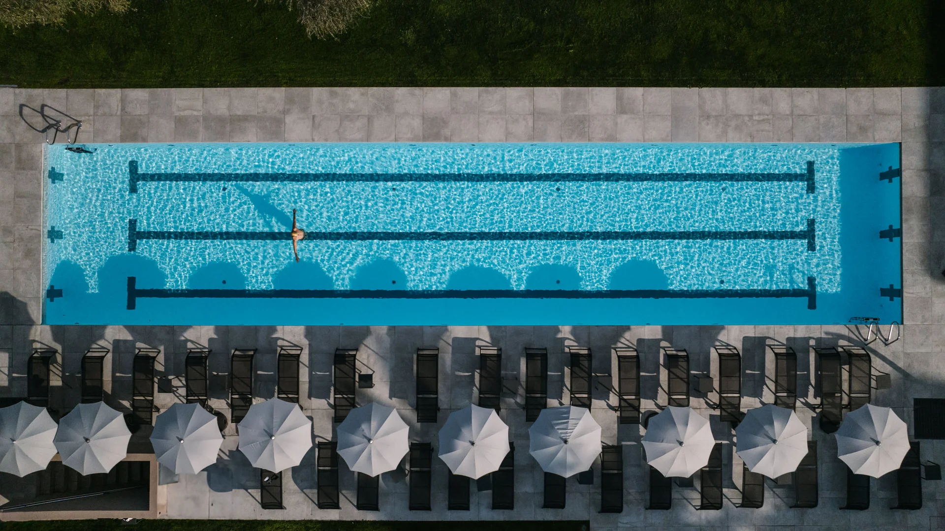Luftaufnahme eines Schwimmbads mit einer Person im Wasser und Liegestühlen mit Sonnenschirmen