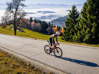 Cyclist rides on mountain road under sunny autumn sky with fog in valley