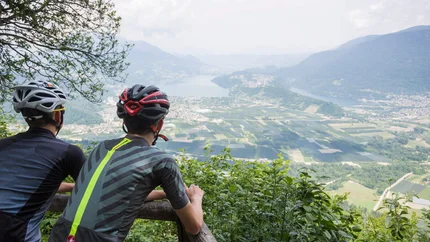 Zwei Radfahrer blicken von einem Berg auf ein Tal mit Fluss und Feldern