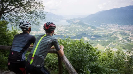 Two cyclists overlooking a valley with river and fields from a mountain