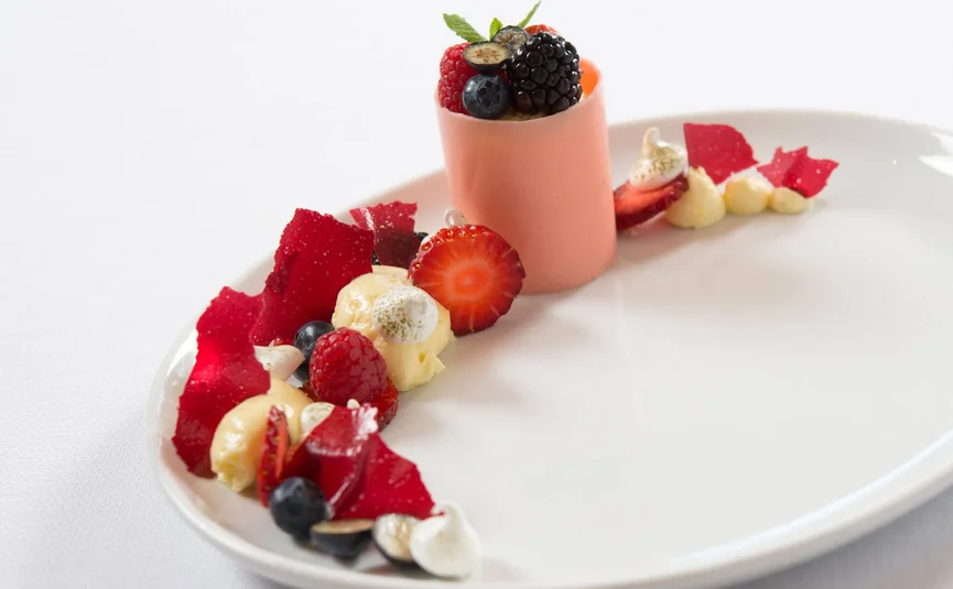 Kunstvoll angerichtetes Dessert mit Beeren, Creme und roten Fruchtblättern auf weißem Teller