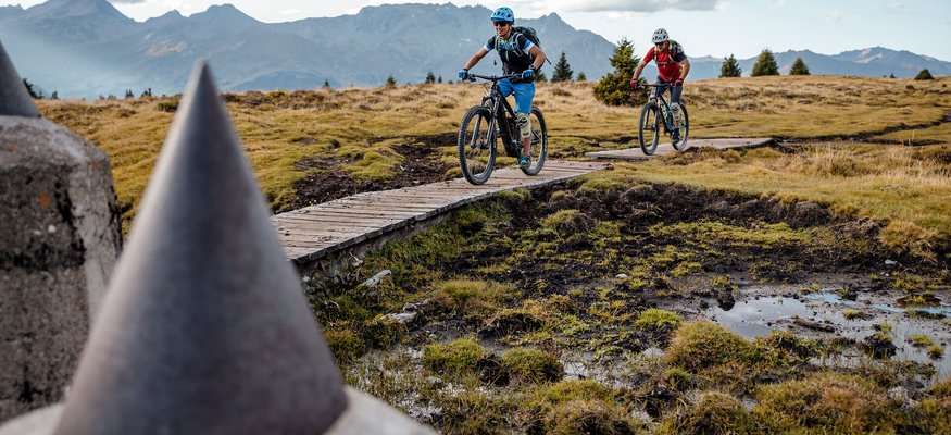 Zwei Mountainbiker fahren auf einem Holzsteg in bergiger Landschaft