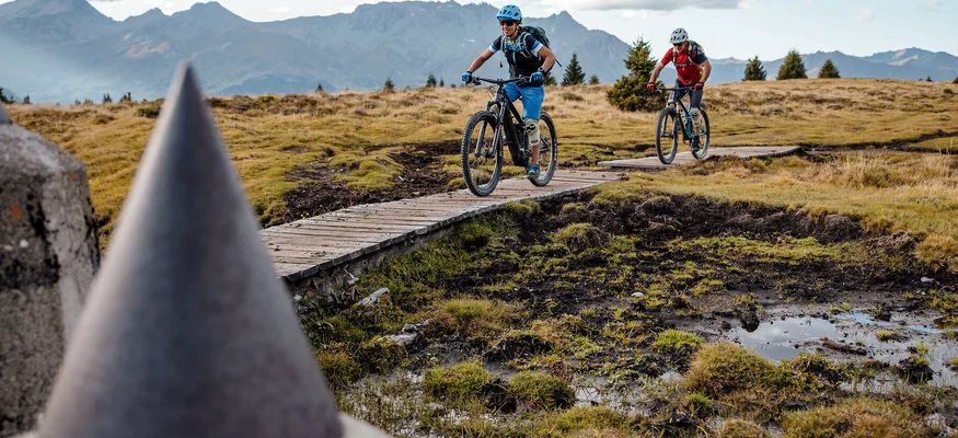 Zwei Mountainbiker fahren auf einem Holzsteg in bergiger Landschaft